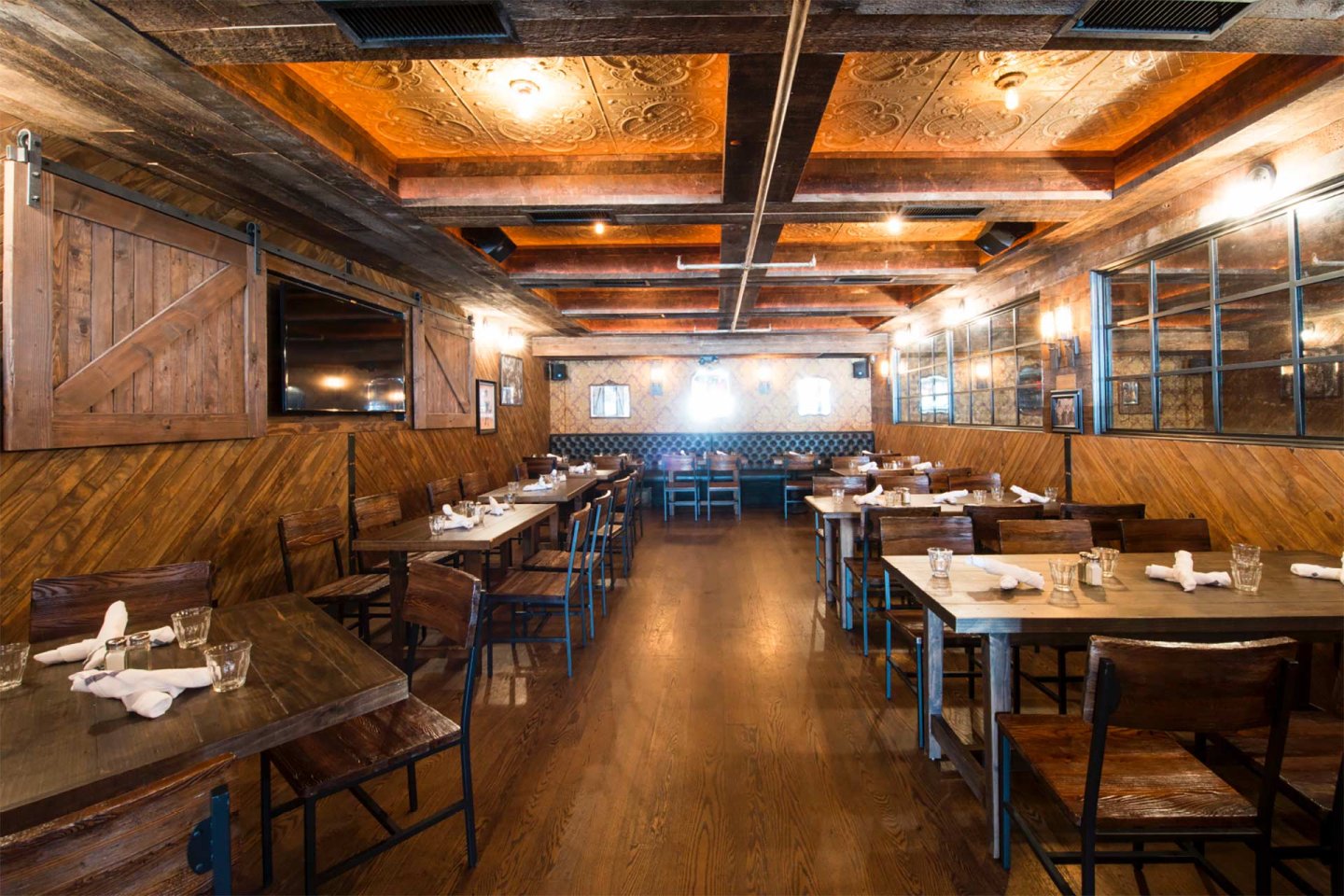 A warmly lit rustic restaurant dining area with wooden floors, tables, and chairs, featuring a decorative ceiling, barn-style doors, and place settings of glasses and napkins on each table.