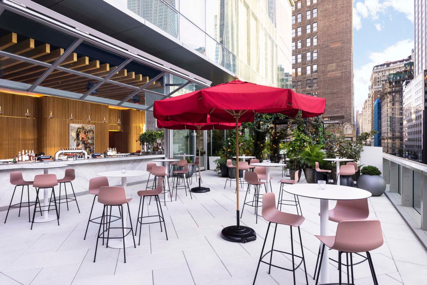 Rooftop patio with high tables and chairs, featuring a large red umbrella. A modern bar area with a wooden facade is in the background. Surrounded by tall buildings, the space is bright and inviting, filled with potted plants and city views.