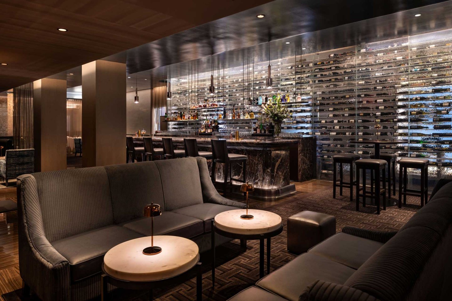 The martini lounge bar interior featuring plush seating arrangements around small tables. A sleek bar counter with high stools is in the background, adorned with various bottles and flowers. The ambiance is luxurious and inviting.