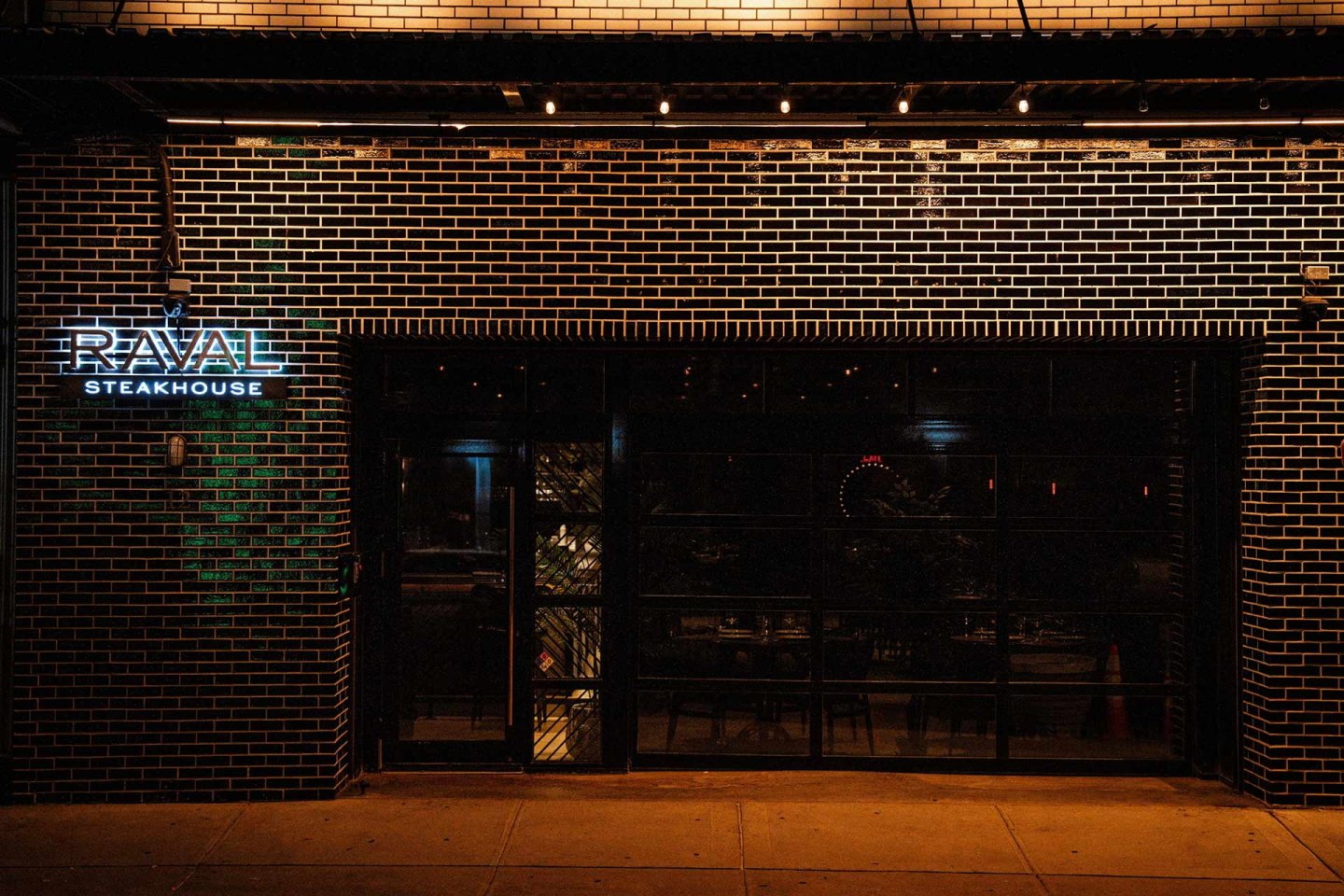 Nighttime view of a brick building with a lit sign reading "RAVAL STEAKHOUSE" on the left, and a large glass door or window revealing a dimly lit interior with tables and chairs inside.
