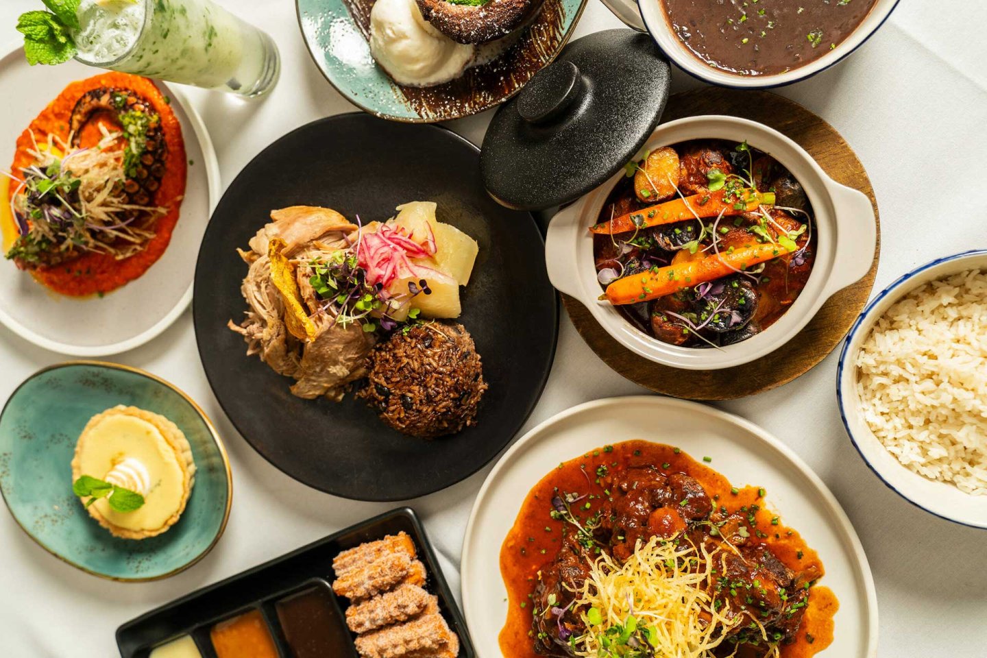 An overhead view of assorted dishes including rice, beef stew, vegetables, dessert, sauces, and a green beverage, all artfully arranged on a white tablecloth.