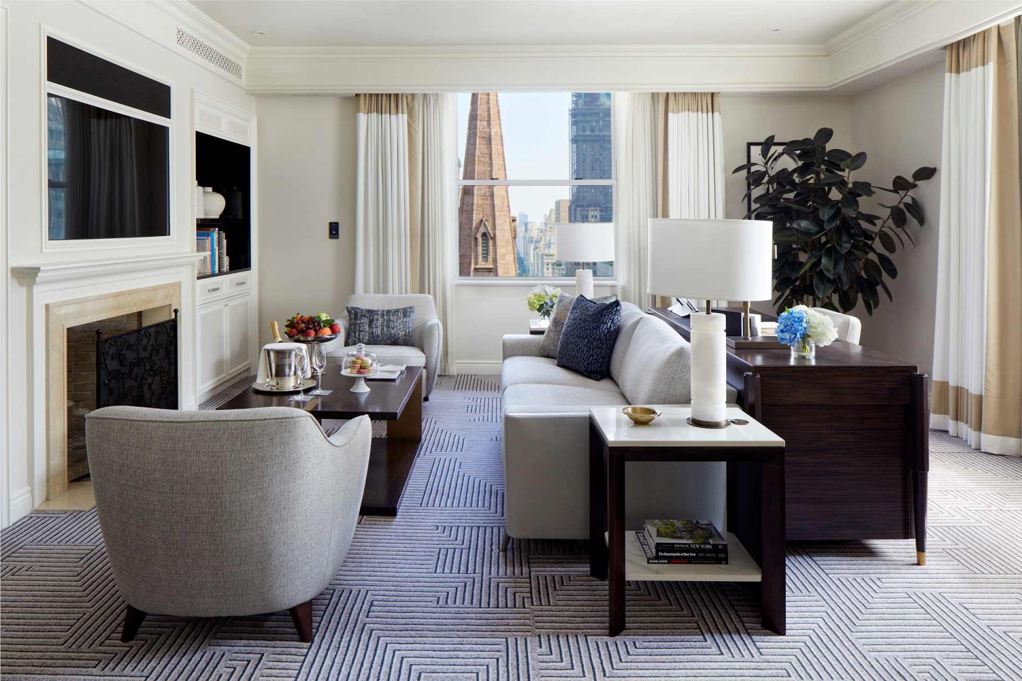 A modern living room with light gray furniture, a dark wooden coffee table, and a side table with books. A large window offers a cityscape view.