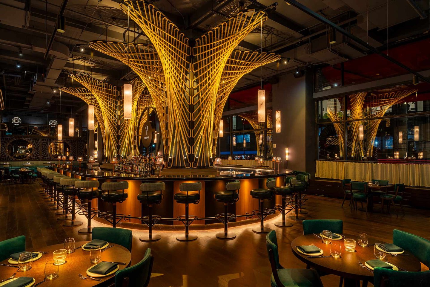 A stylish, modern restaurant interior with dramatic golden light fixtures resembling large, woven sculptures above a circular bar, surrounded by green velvet chairs and set tables under warm lighting.