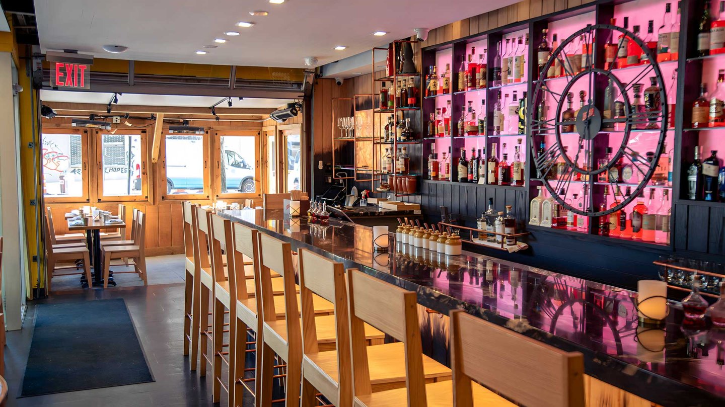 A modern bar with wooden chairs lined along a shiny counter, shelves of liquor bottles against a pink-lit wall, and large windows letting in natural light.