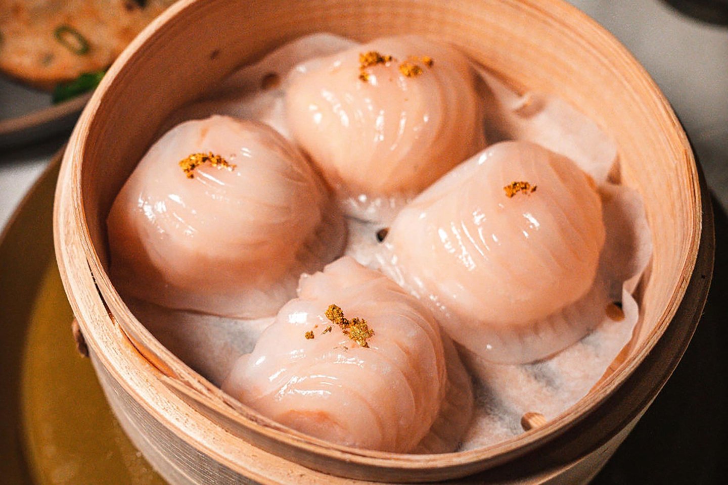 Four translucent shrimp dumplings topped with a small amount of seasoning are arranged in a bamboo steamer lined with parchment paper.
