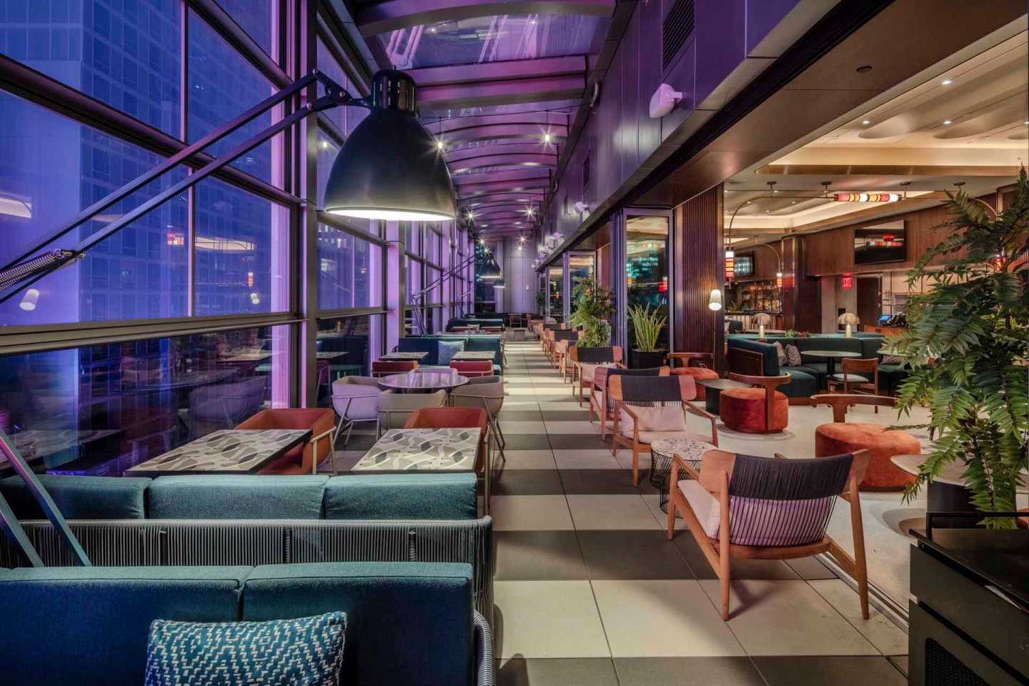 Stylish indoor lounge with modern decor featuring blue-lit windows on the left, comfortable seating in various colors, tables, and a large overhead lamp. The space is adorned with plants and has a sophisticated, relaxed ambiance.