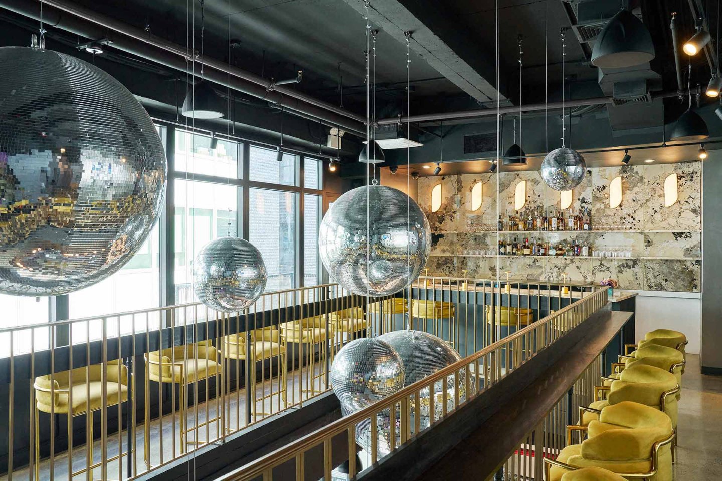 Modern bar interior with large disco balls hanging from the ceiling, yellow velvet bar stools, marble bar counter, and shelves stocked with bottles, all set against large windows and stylish lighting.