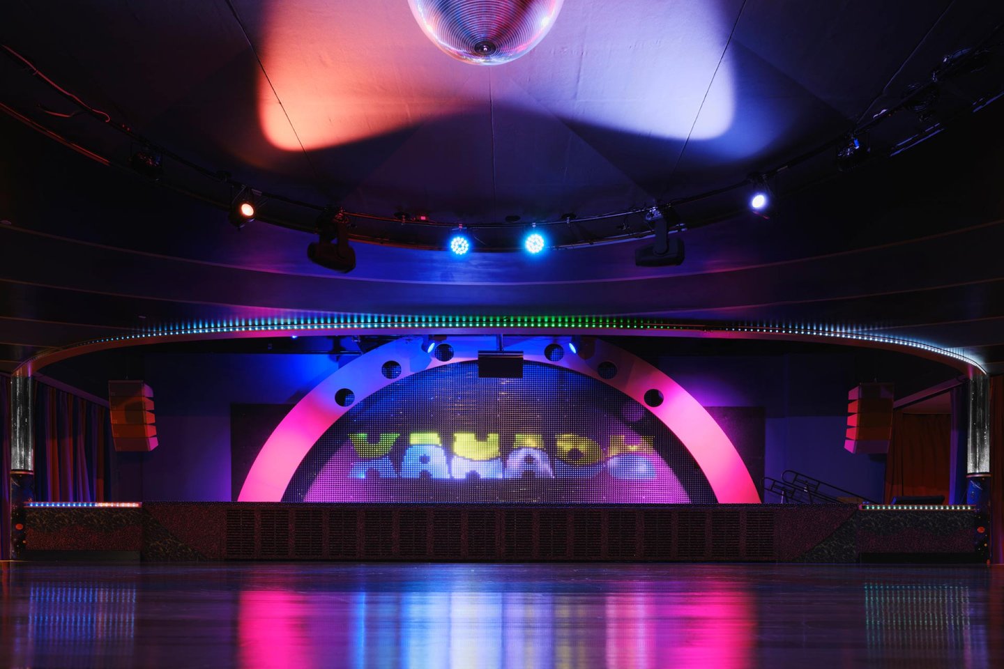 A colorful, empty stage with a disco ball overhead, vibrant spotlights, and the word "XANADU" illuminated on the back wall, reflecting onto a polished floor in a modern performance venue.