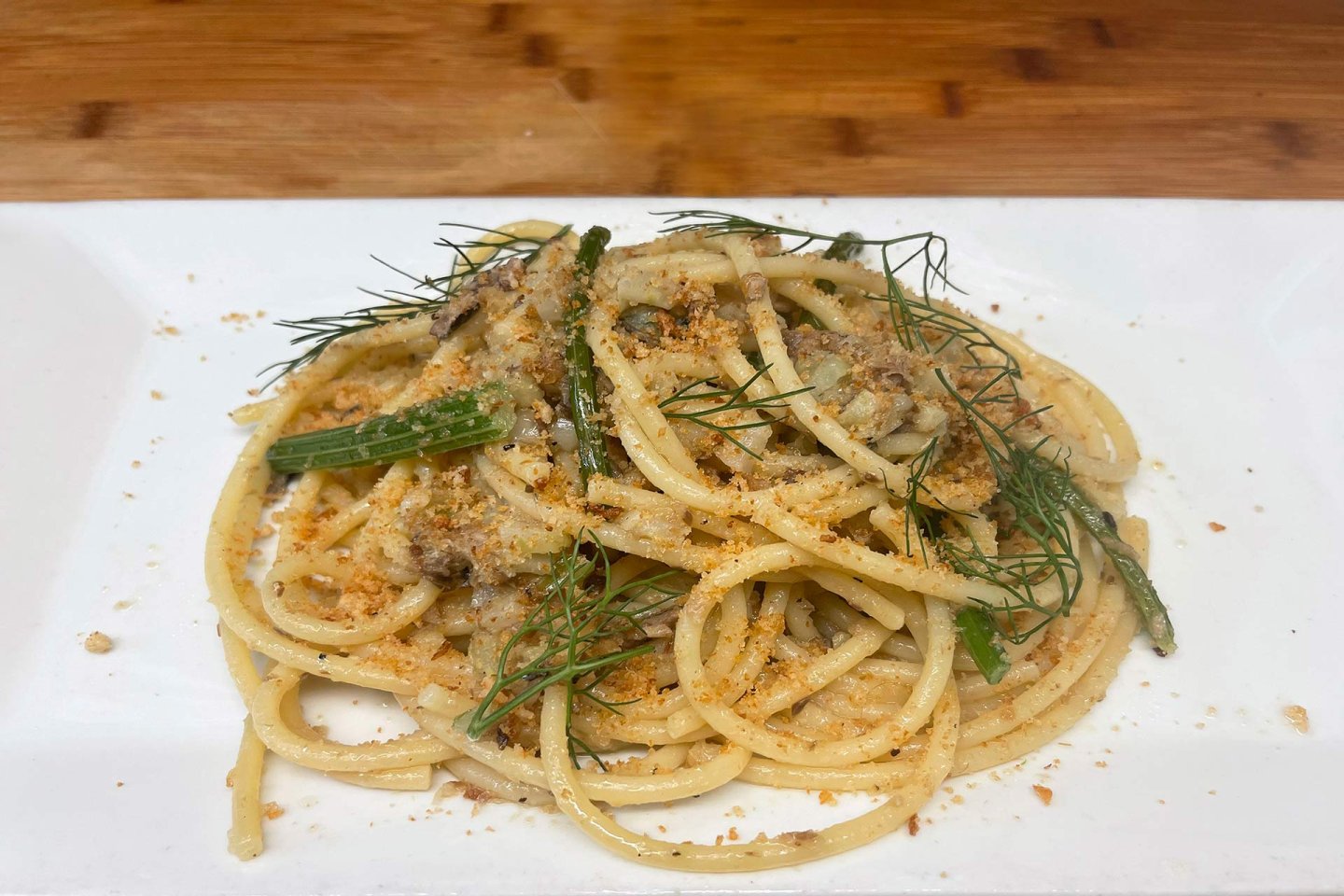 A plate of pasta with sausage, green beans, and herbs, garnished with fresh dill and breadcrumbs, served on a white rectangular dish on a wooden table.