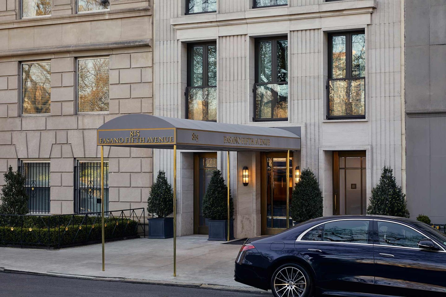 The entrance of a building with a gray canopy labeled "85 East 80th / 85 East 80th Avenue," flanked by potted plants. A black luxury car is parked on the street in front.