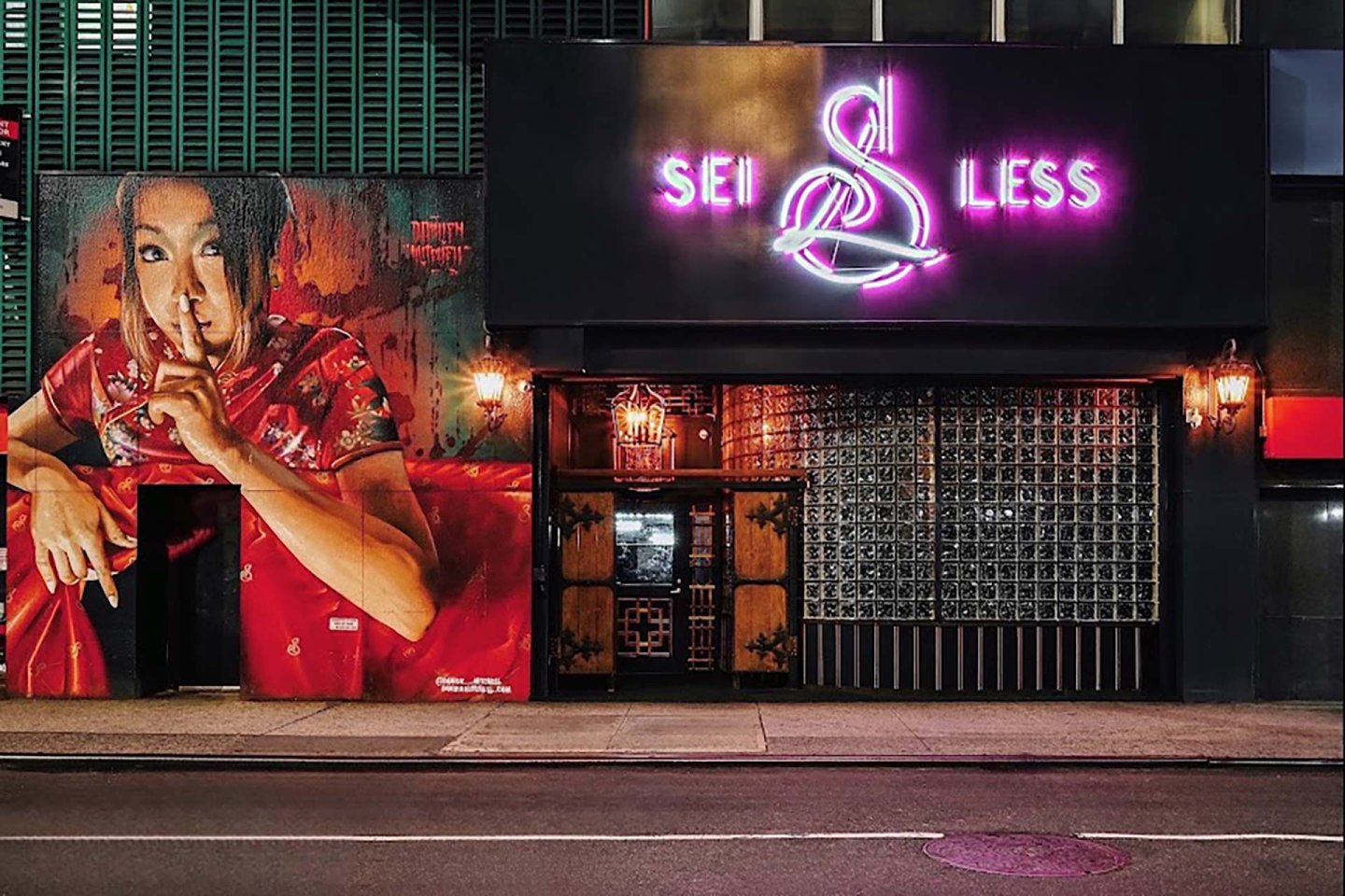 A street view of a building at night with a large mural of a woman in a red dress holding a finger to her lips, next to a neon sign reading "SEI LESS" above a black entrance.