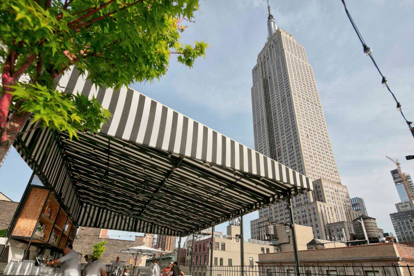 A black-and-white striped canopy covers a rooftop bar with people sitting beneath it. In the background, the tall Empire State Building rises against a blue sky in New York City. Tree branches frame the left side of the image.