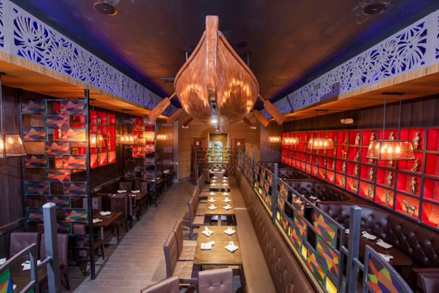 Restaurant interior with a nautical theme, featuring a large wooden boat hanging from the ceiling. Dark leather seating lines both sides of long wooden tables. Warm lighting from hanging fixtures and vibrant red and blue wall art create a cozy atmosphere.
