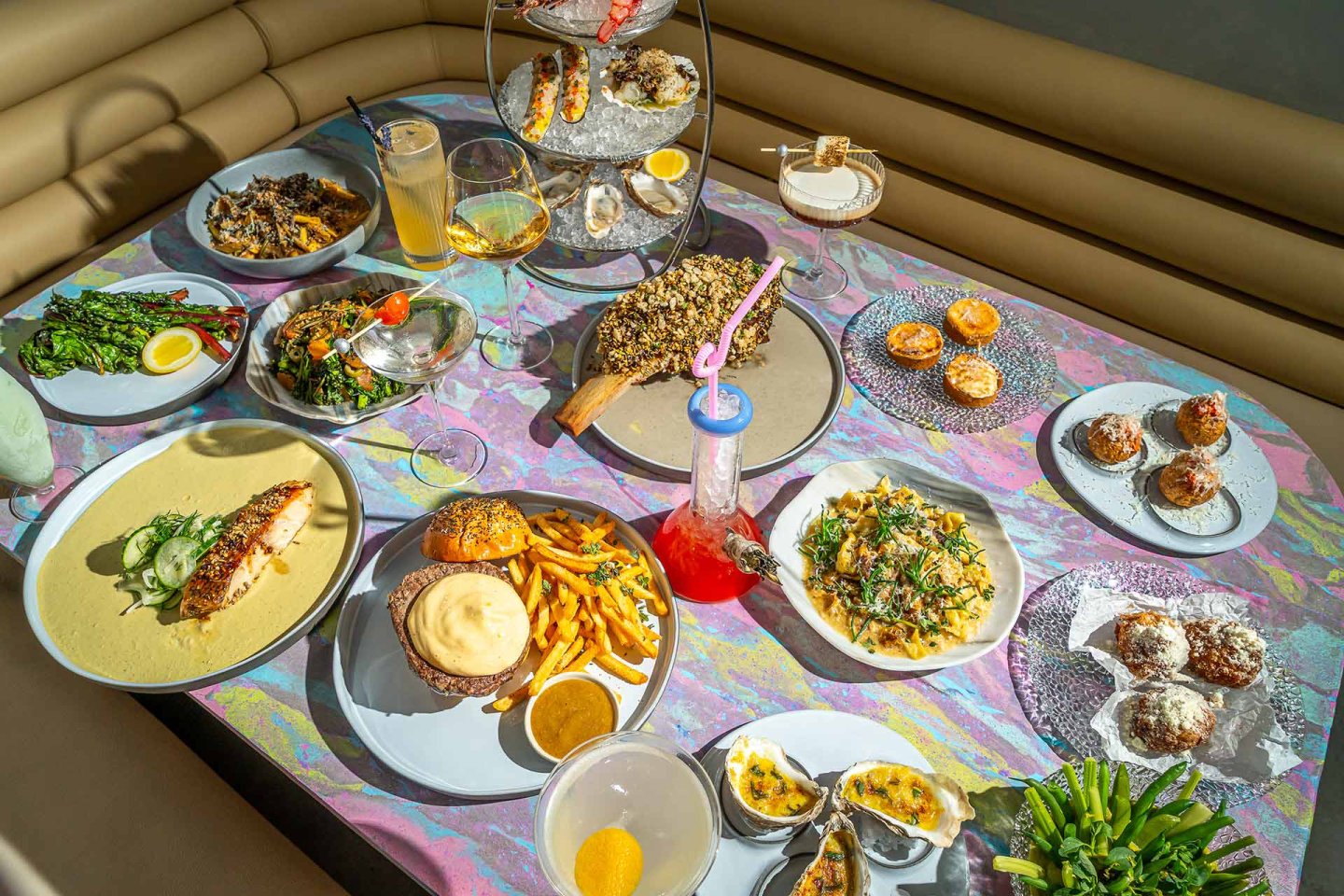 A table with a variety of colorful dishes, including seafood, fries, salad, pastries, drinks, and desserts, set on a pastel marble-patterned tablecloth in a modern dining setting.