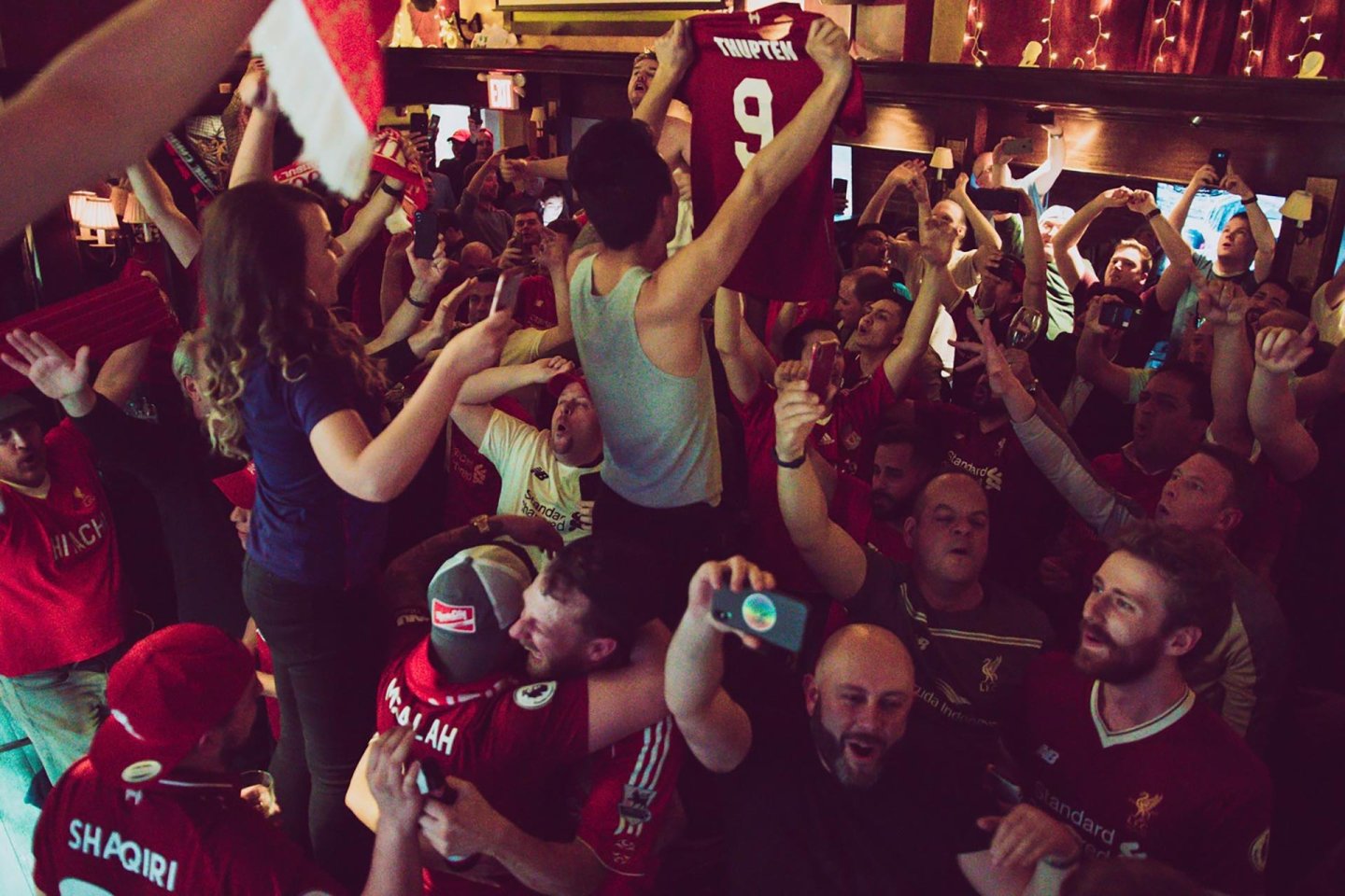 A lively crowd of soccer fans, many wearing red jerseys, cheer and celebrate together in a dimly lit pub. One person holds up a jersey with the name “Firmino” and number 9 as others capture the moment on their phones.