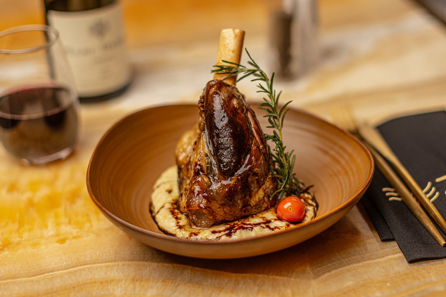 A braised lamb shank served on creamy mashed potatoes, garnished with a cherry tomato, rosemary sprig, and a drizzle of sauce, with a glass of red wine and utensils on the side.