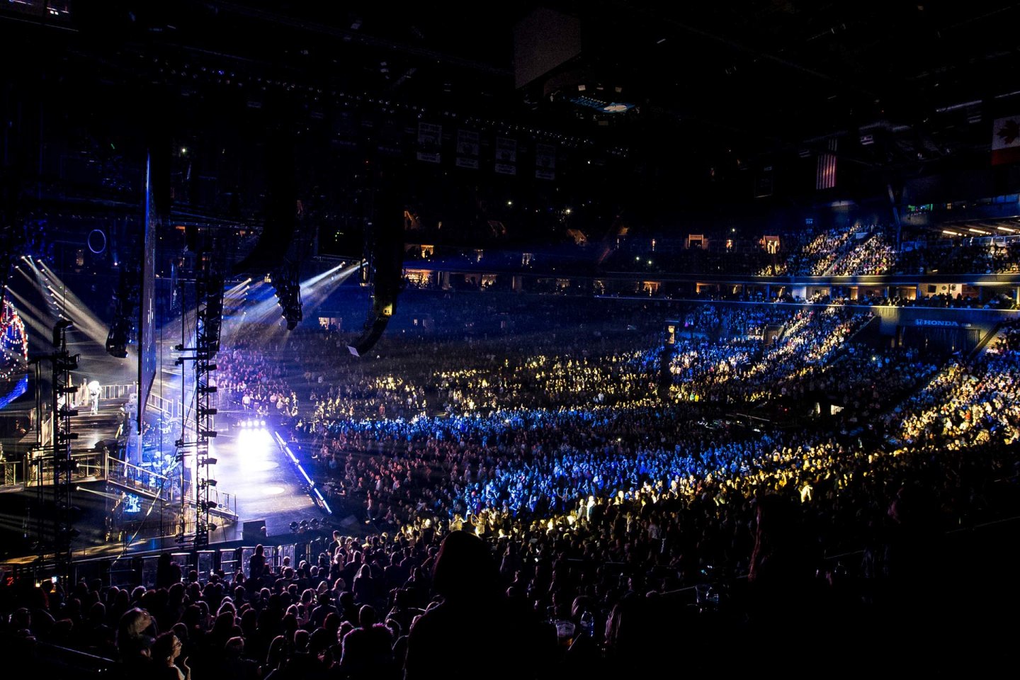 A large, packed indoor arena glows with stage lights during a concert, illuminating a cheering crowd and performers on a brightly lit stage. Blue and yellow lighting highlights sections of the audience.