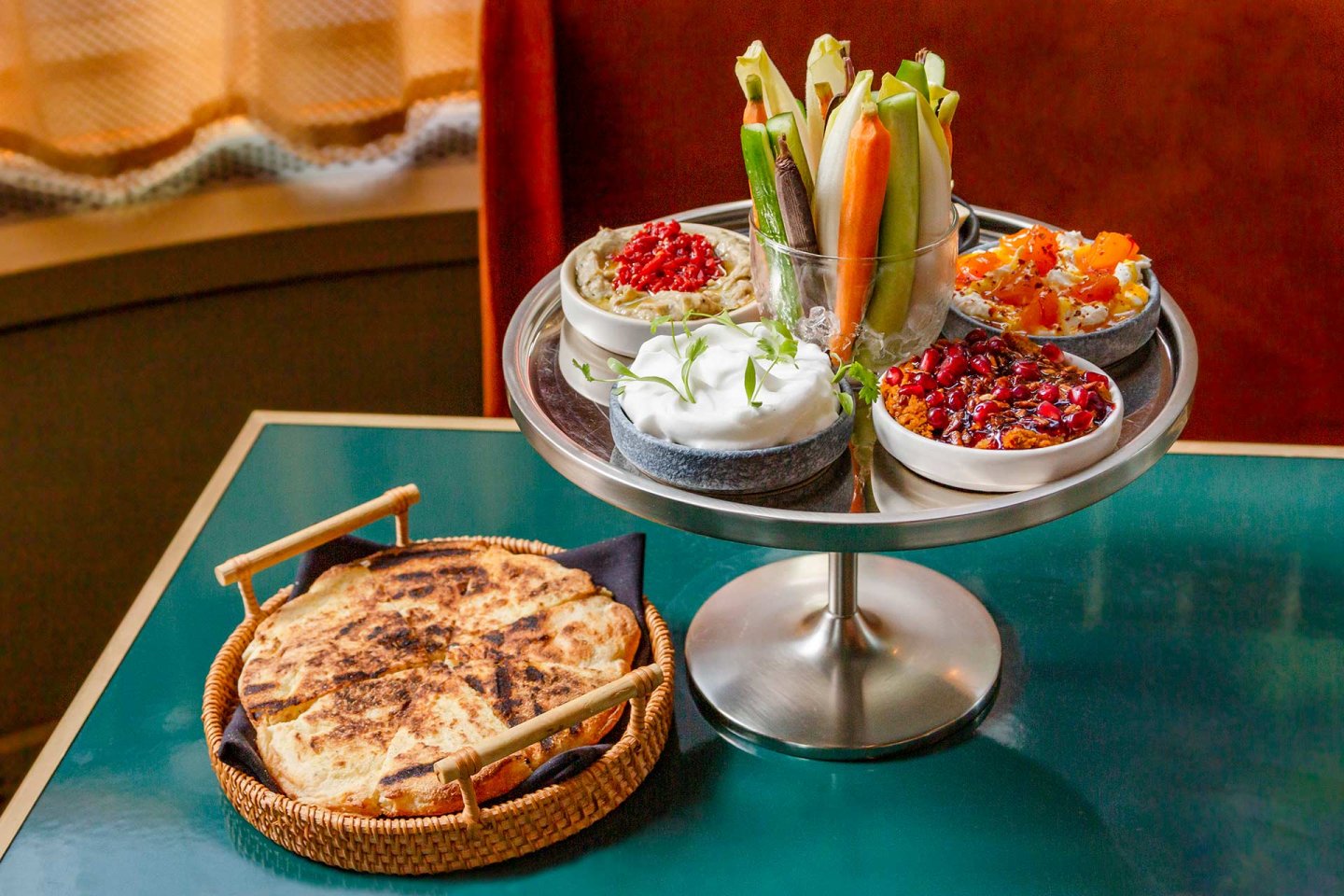 A tiered tray displays assorted vegetable sticks and five bowls of colorful dips and spreads, alongside a basket with grilled flatbread on a green table.