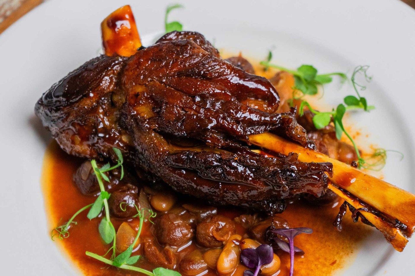 A glazed braised lamb shank served on a white plate with a rich brown sauce, accompanied by beans and garnished with fresh green herbs.