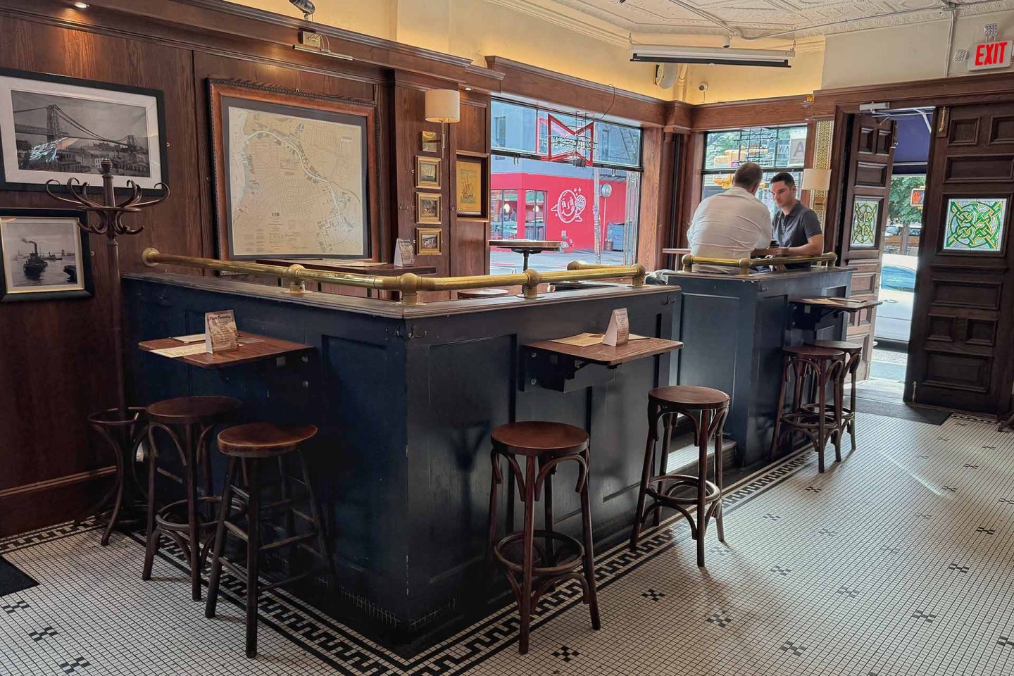 A cozy bar with wooden stools and a black counter, map and photos on the walls, tiled floor, and two people talking by the window. Sunlight streams in from outside where a red sign is visible.