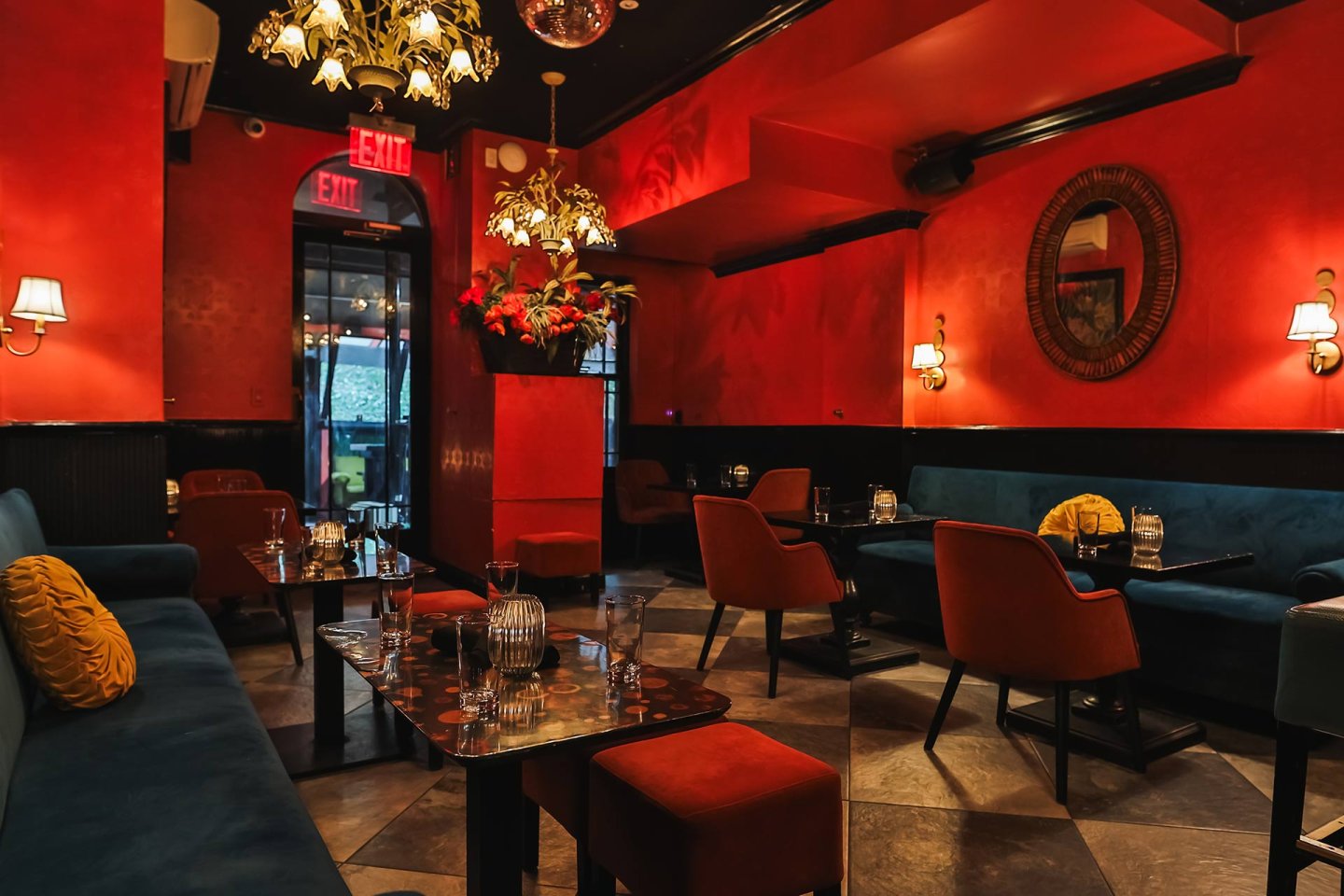 Stylish restaurant interior with vibrant red walls, modern chandeliers, dark blue sofas, orange chairs, elegant table settings, wall art, and a large floral arrangement near the entrance.