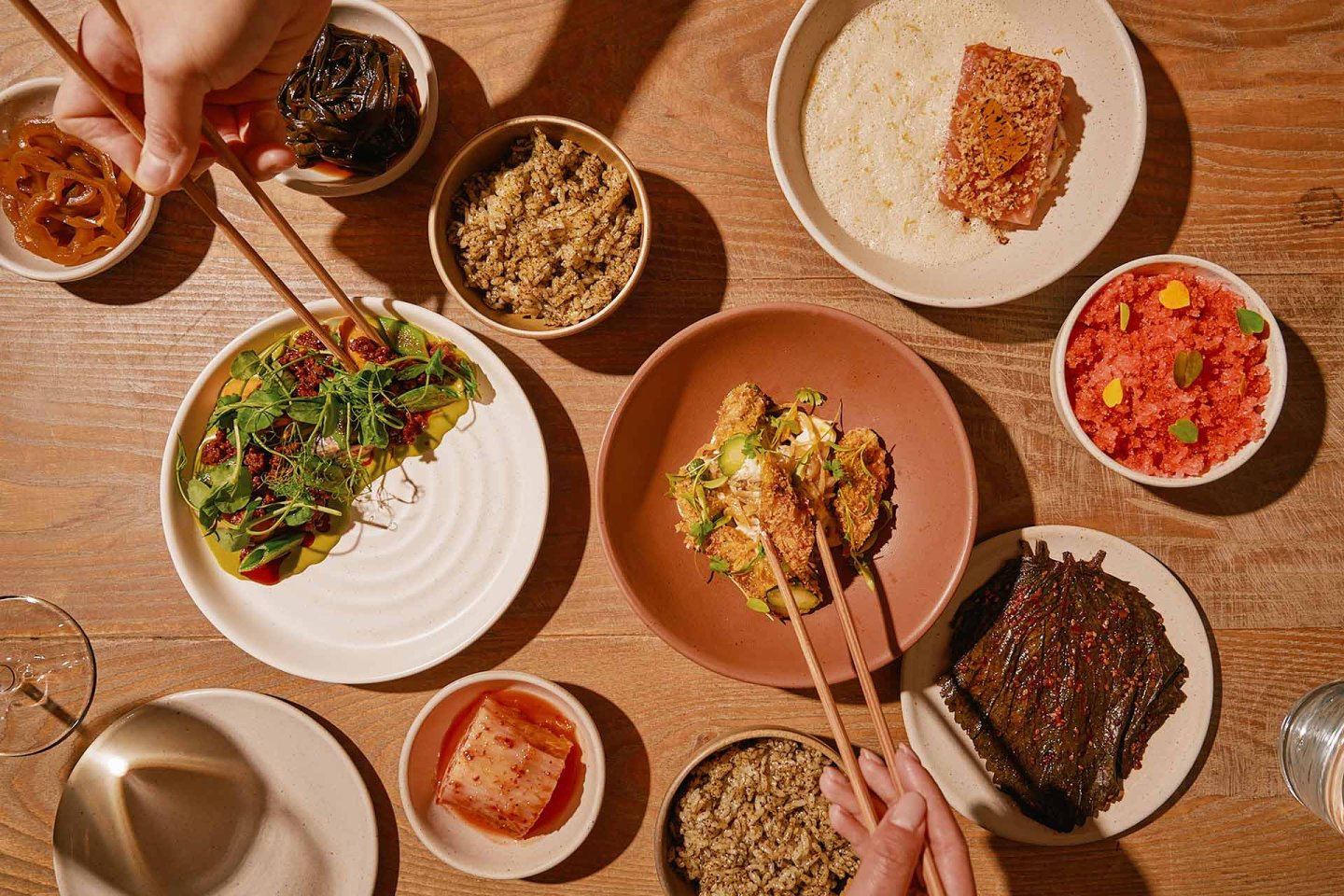 Overhead view of a wooden table set with various small dishes of food, including rice, vegetables, pickles, and fish. Two pairs of hands use chopsticks to pick up food from different plates.