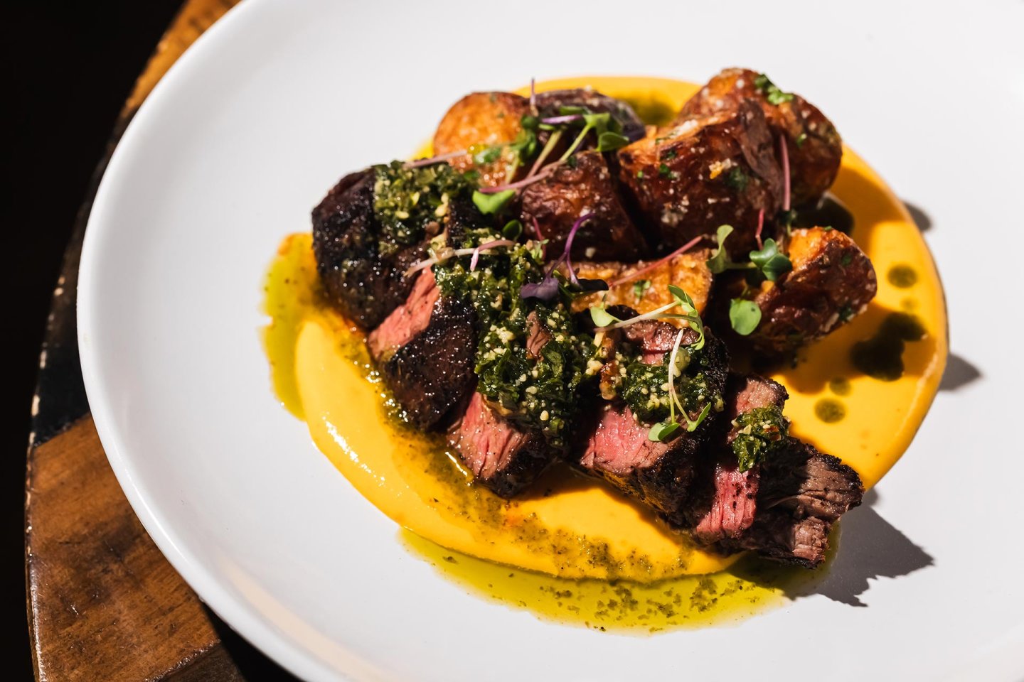 A plate with sliced medium-rare steak topped with green herb sauce, served on a bed of yellow puree, accompanied by roasted potatoes and garnished with microgreens.