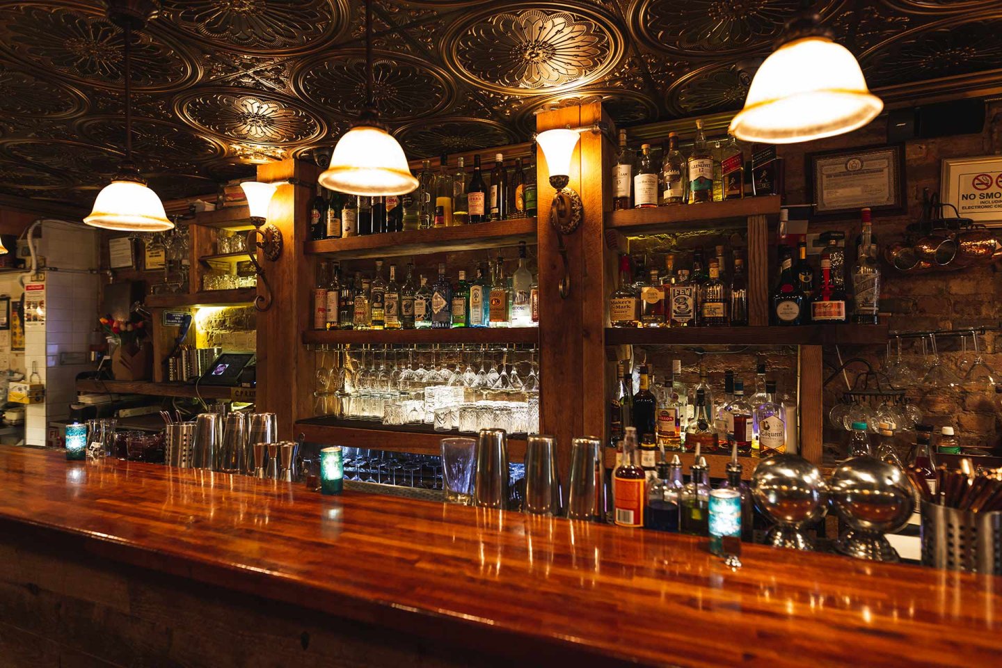 A cozy bar with a polished wooden counter, various liquor bottles and glasses displayed on shelves, warm overhead lights, and vintage-style ceiling tiles, creating an inviting, intimate atmosphere.