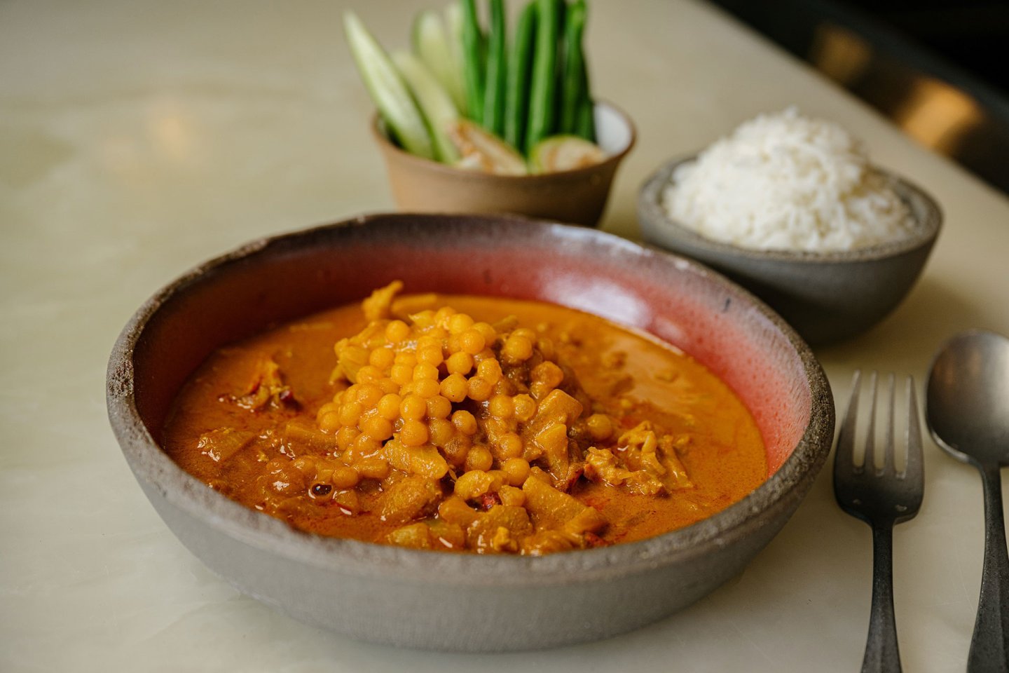 A bowl of curry with chickpeas and meat, served with a side of steamed white rice, fresh vegetables, and utensils on a light-colored table.