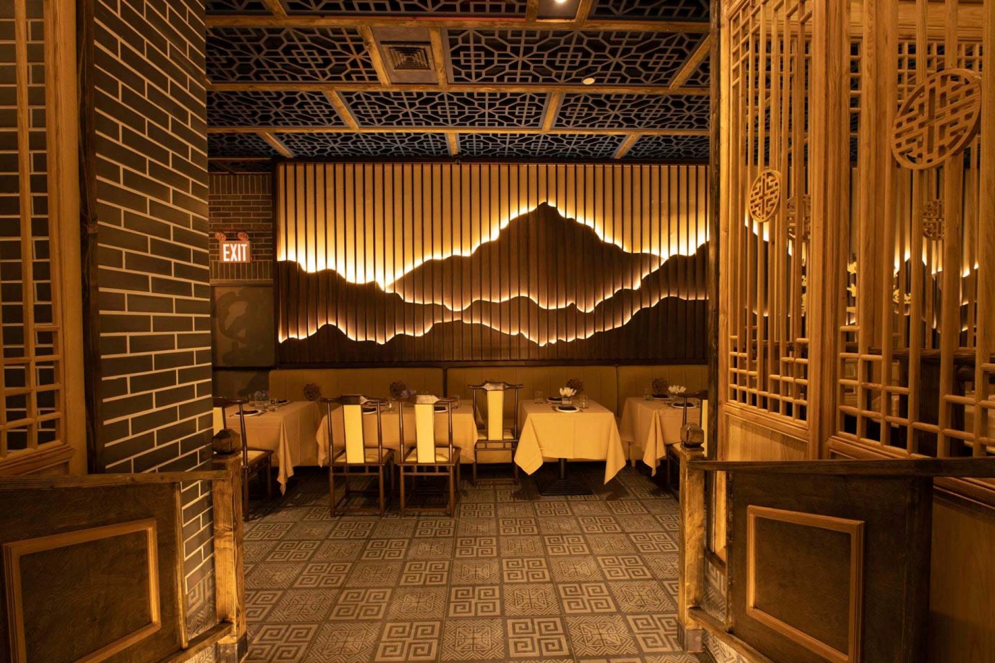 A warmly lit dining area with a mountain silhouette decor on the wall. The room features ornate wooden panels and patterned flooring, with tables set with white tablecloths and elegant tableware. An exit sign is visible in the background.