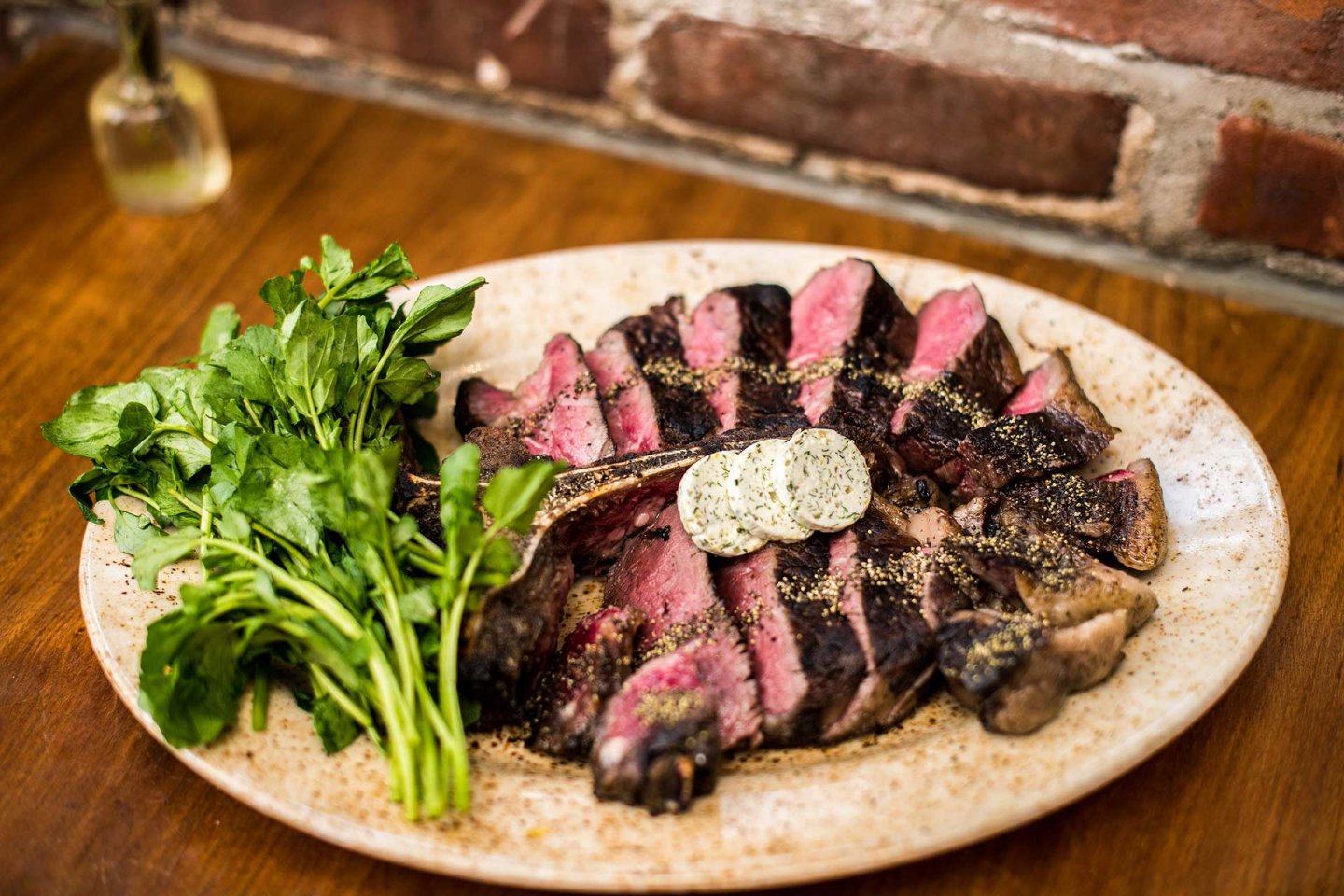 A plate of sliced, medium-rare steak with cracked pepper, topped with herb butter, served next to a bunch of fresh parsley on a rustic plate, set on a wooden table with a brick wall in the background.