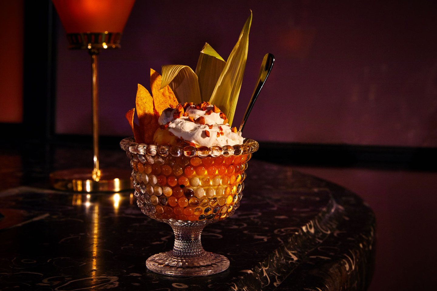 A decorative glass dessert bowl filled with colorful tapioca pearls, whipped cream, fruit slices, and corn leaves sits on a dark marble table, with a spoon and a lamp in the dimly lit background.