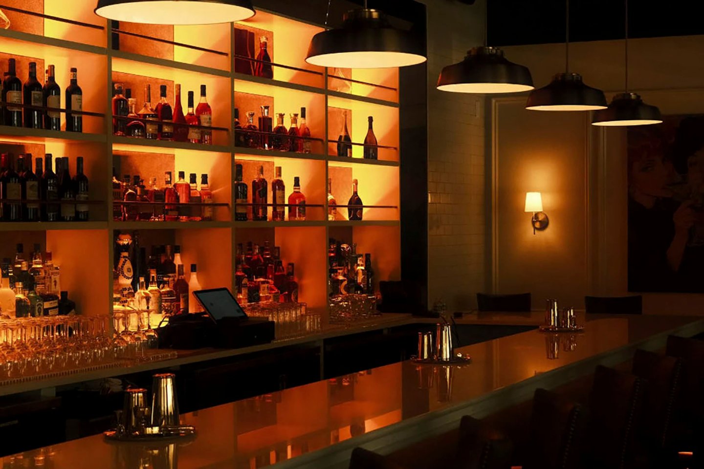 Dimly lit bar with illuminated shelves displaying various bottles of alcohol. A counter with bar stools faces the shelves. Hanging lights create a warm ambience. A painting and wall sconce can be seen on the right.