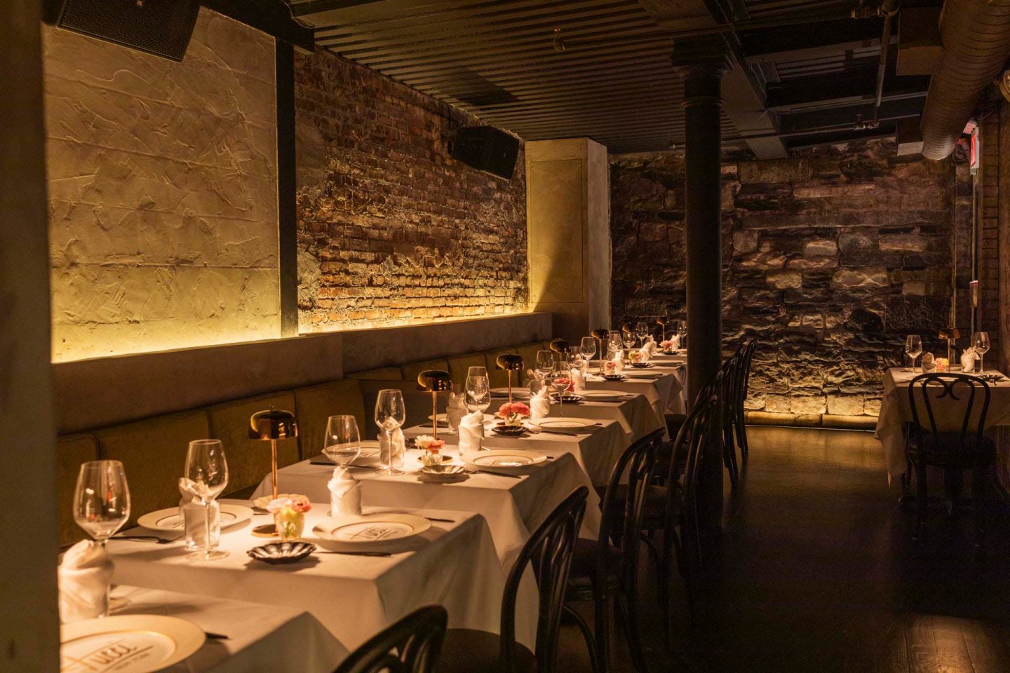 A dimly lit restaurant with exposed brick walls and elegant table settings. Each table has white tablecloths, wine glasses, and small flower arrangements. Black chairs line the tables, creating a cozy and intimate dining atmosphere.