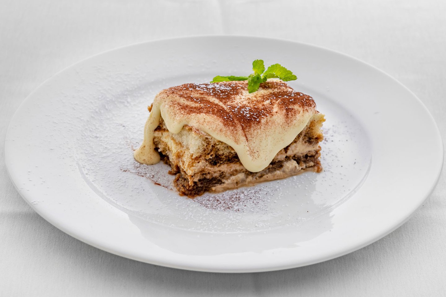 A slice of tiramisu topped with cocoa powder and a mint leaf sits on a white plate sprinkled with powdered sugar, set against a white background.