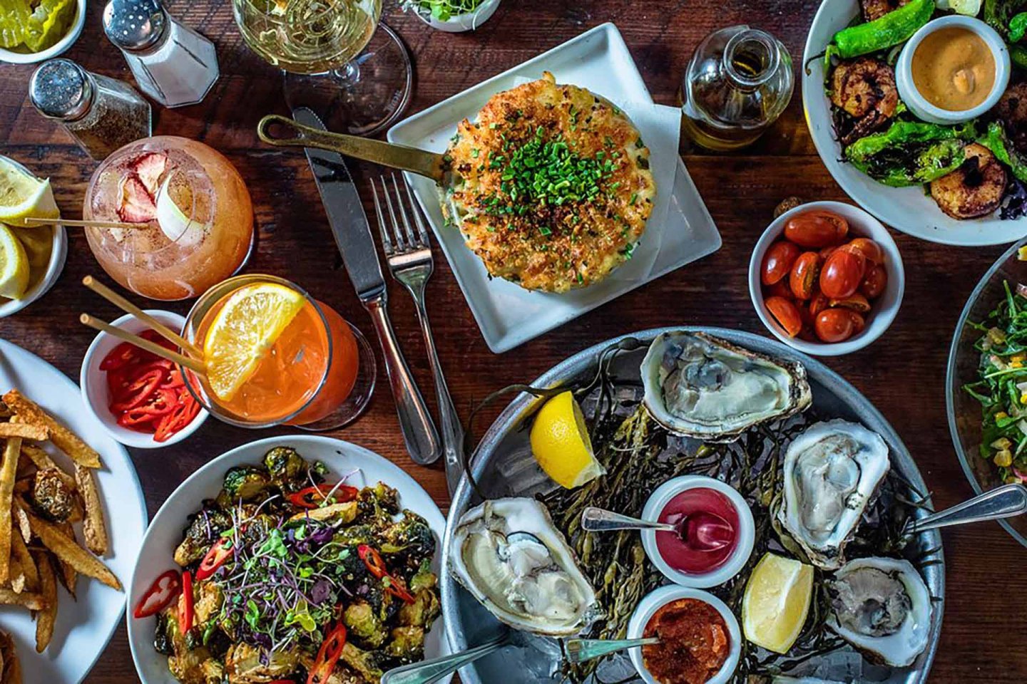 A table filled with various dishes including oysters on ice with lemon, a baked dish topped with herbs, fried foods, cherry tomatoes, cocktails, and glasses of white wine, all arranged on a wooden surface.