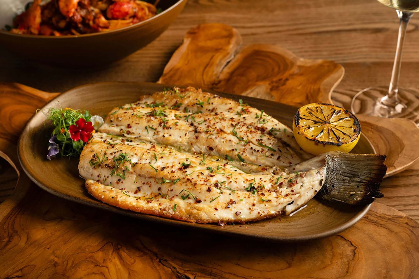 Grilled whole fish served on a brown plate with herbs, a charred lemon half, and a small garnish; wooden table and a glass of white wine in the background.