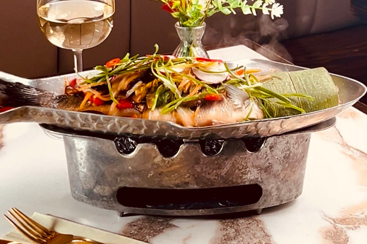 A whole steamed fish topped with sliced vegetables and herbs is served on a metal platter over a heated base, next to a glass of white wine and a small flower vase on a marble table.