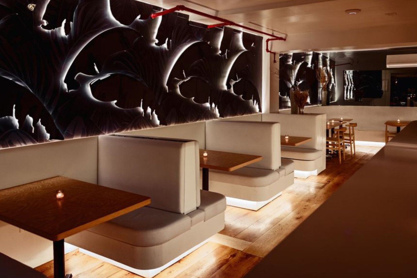 Modern restaurant interior with cushioned booth seating, wooden tables, and ambient lighting. Dark, abstract wall art and a wooden floor create a cozy, upscale atmosphere. A mirror reflects part of the space.
