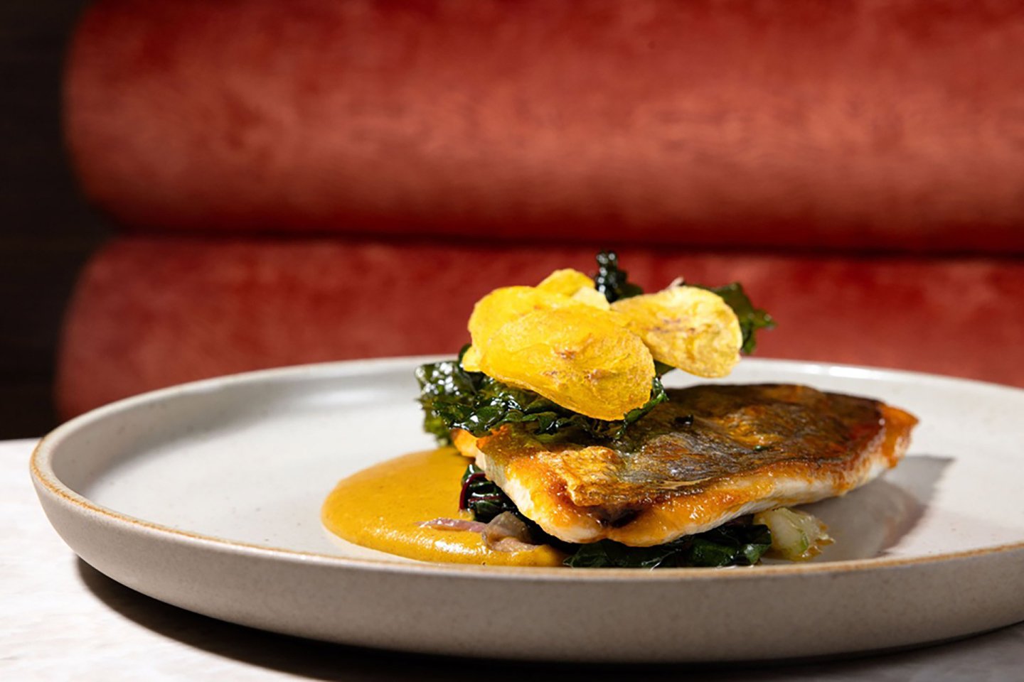 A gourmet dish featuring a seared fish fillet with crispy skin, served on a creamy orange sauce, topped with kale and thin yellow crisps, presented on a gray plate against a red cushioned background.