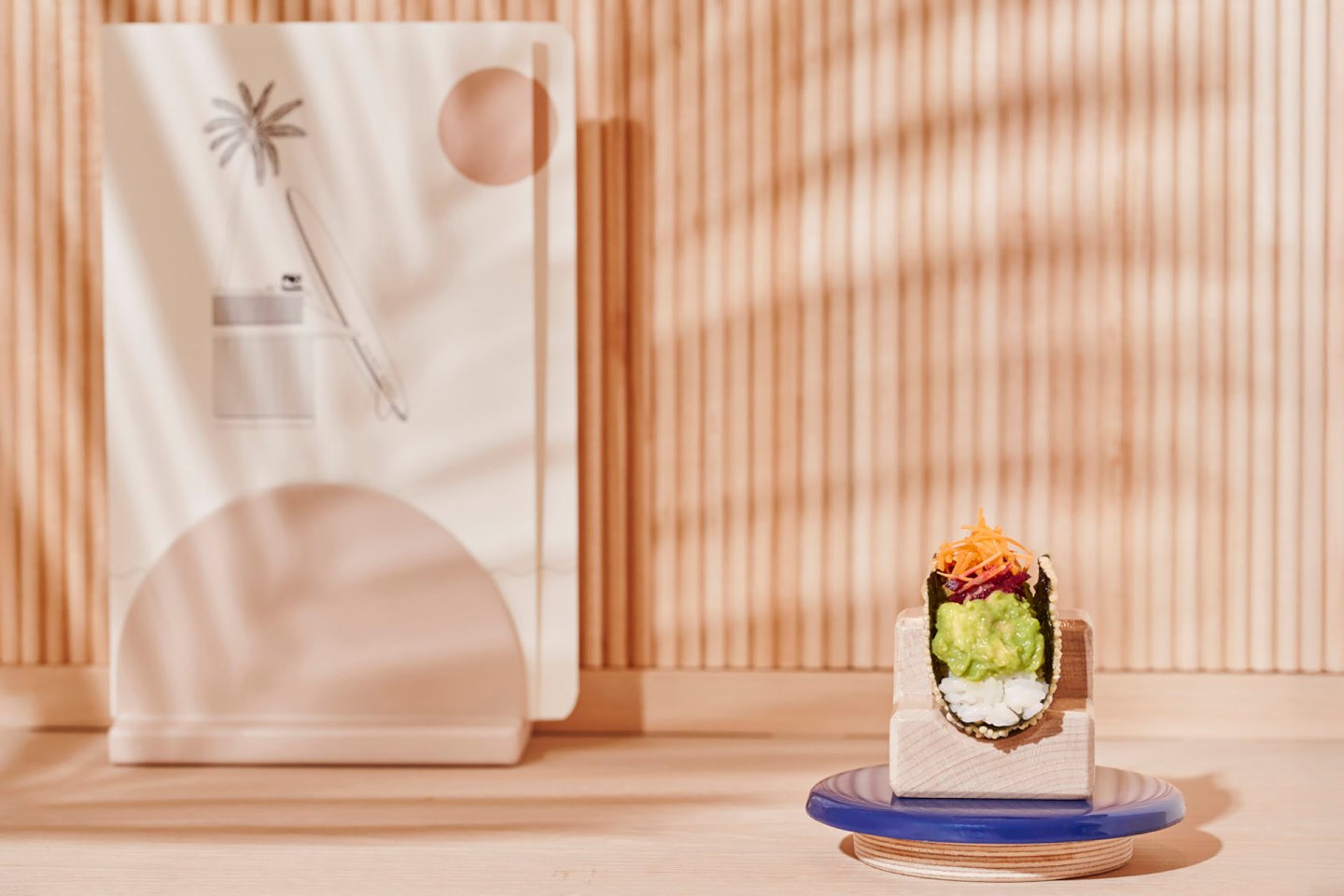 A sushi roll wrapped in seaweed, served on a wooden block with blue and white plates, sits on a wooden surface. Behind it is a minimalist artwork with abstract shapes and a palm tree, with striped shadows on a wooden background.