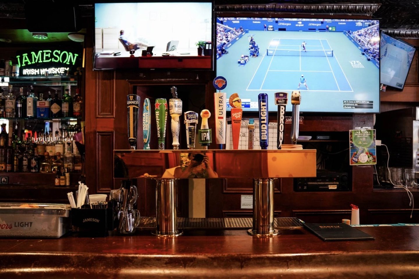 A bar counter with beer taps in front of shelves of liquor bottles. Two TVs are above the bar, one showing a tennis match and the other displaying a scene with a person at a desk. A green Jameson sign is on the left.