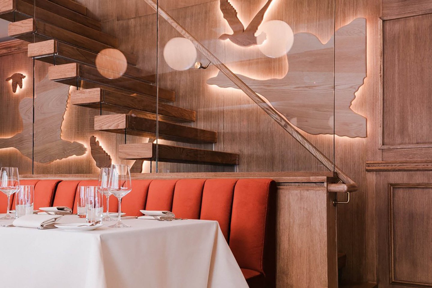 Elegant restaurant interior with cream-colored chairs, tables set with white tablecloths and glassware, large windows letting in natural light, and a floral arrangement on a wooden sideboard.