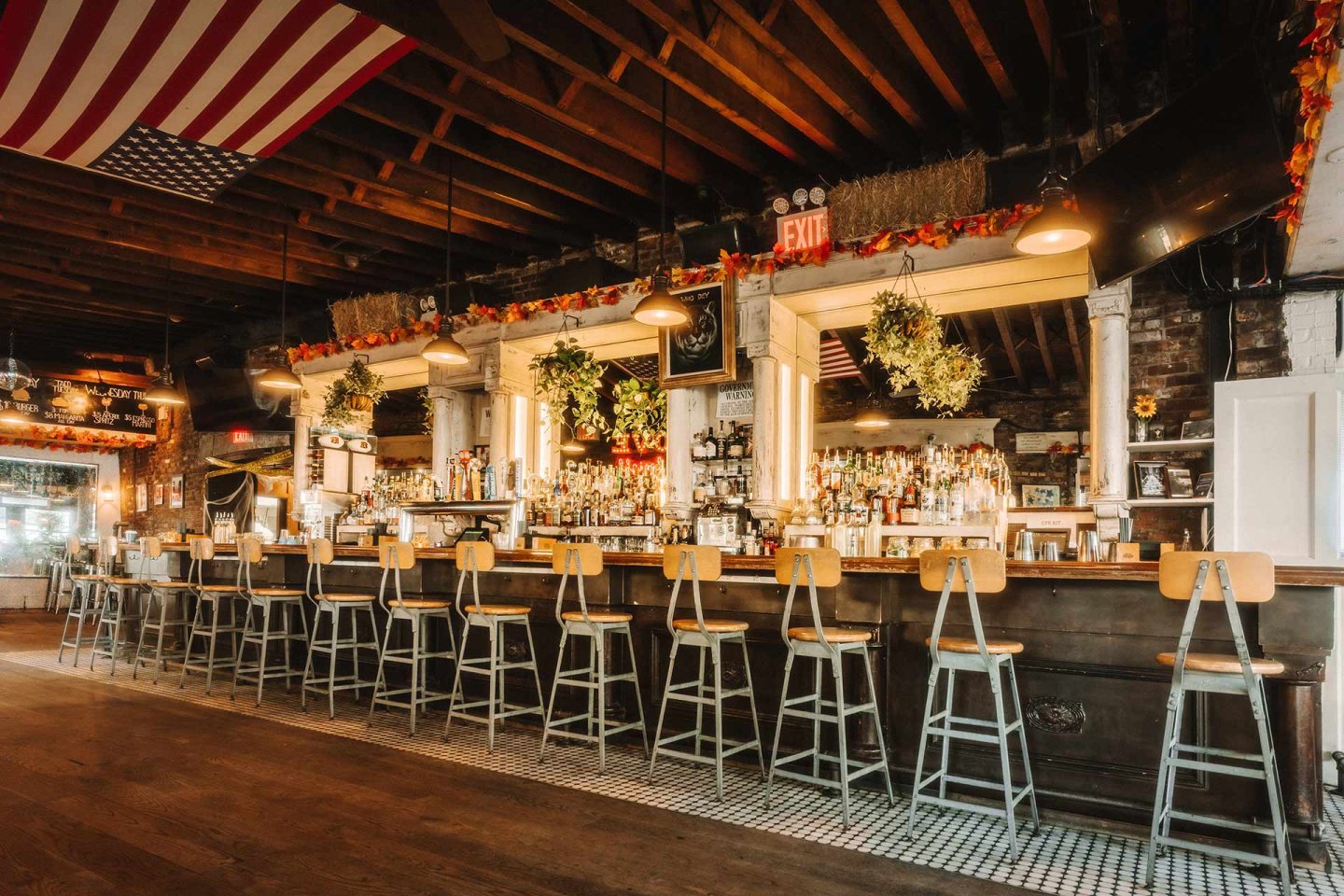 A rustic bar with a long wooden counter, metal barstools, hanging plants, shelves of bottles, exposed beams, and an American flag hanging from the ceiling. Warm lighting and autumn decorations create a cozy atmosphere.