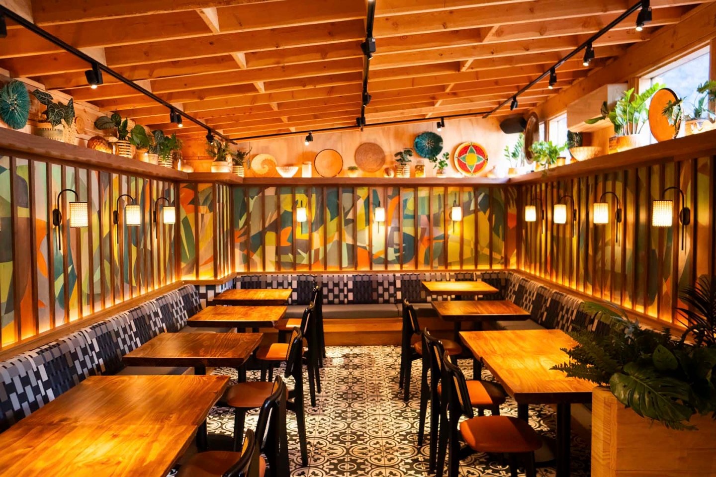 A cozy restaurant interior with wooden tables and chairs, vibrant patterned walls, and warm hanging lights. The ceiling has exposed beams, and plants decorate the high shelves, complementing the artistic atmosphere.