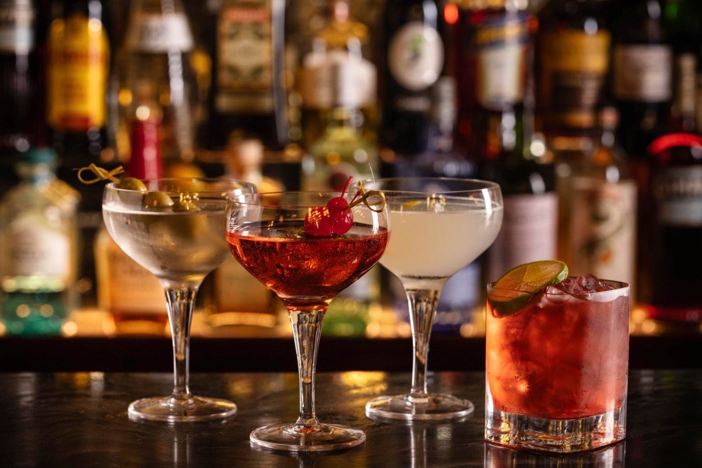 Four cocktails in elegant glasses are lined up on a bar counter. They include a martini with olives, a red drink with cherries, a clear drink with an olive, and a pink cocktail with a lime wedge. Bottles are blurred in the background.