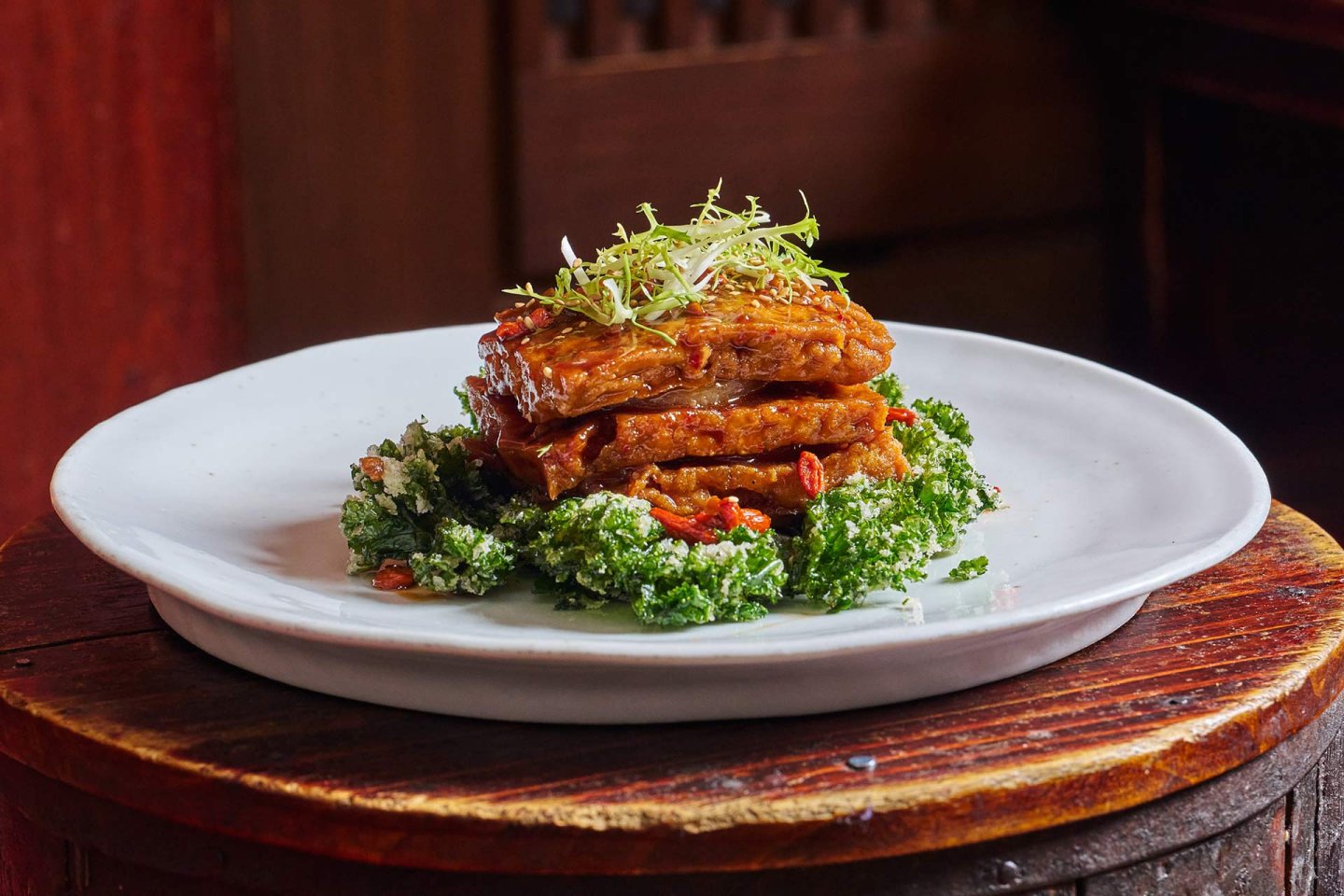 A stack of glazed tofu slices is served on a bed of sautéed kale and garnished with fresh microgreens, presented on a white plate atop a rustic wooden surface.