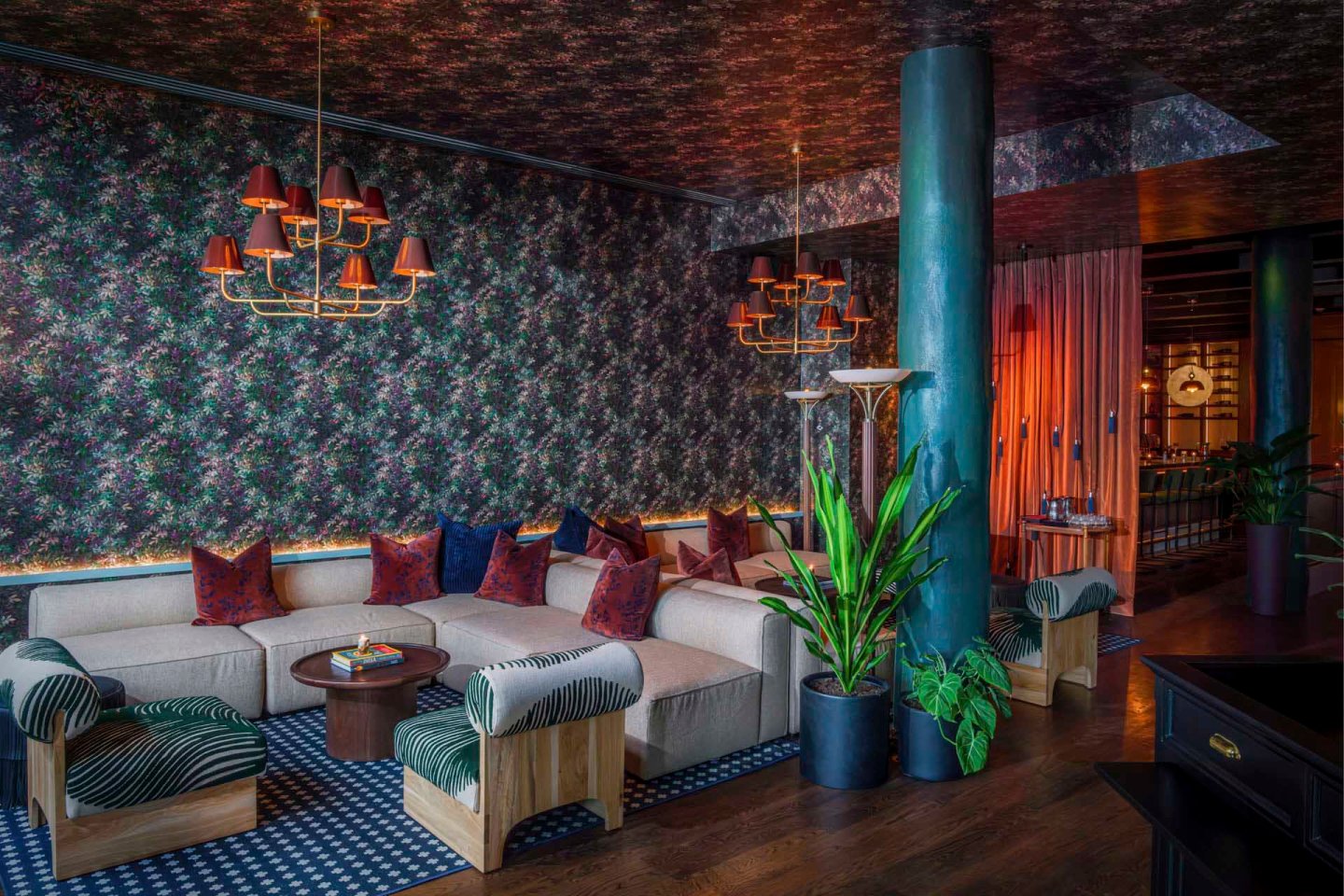 A stylish living room with a U-shaped beige sofa adorned with red and blue pillows, two striped ottomans, a round wooden coffee table, floral-patterned walls, a chandelier, and potted plants. Warm lighting enhances the cozy atmosphere.