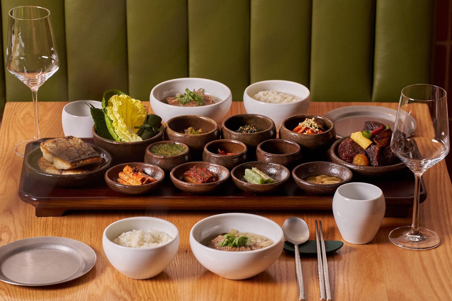 A wooden table set with an assortment of Korean dishes in small brown bowls, including rice, vegetables, meats, and sauces, accompanied by two wine glasses, metal chopsticks, and ceramic cups.