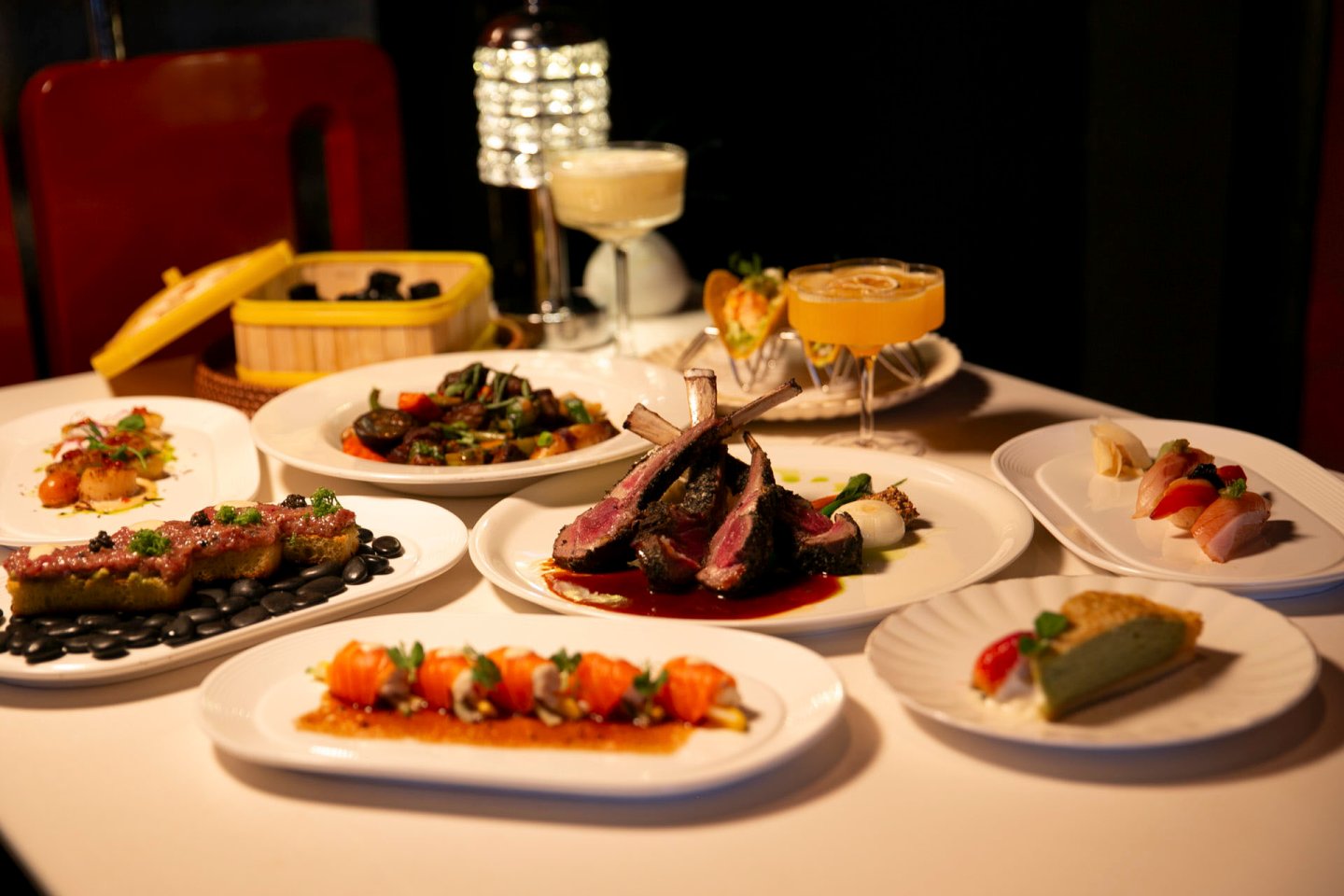 A table set with various plates of gourmet food, including lamb chops, sushi, and assorted appetizers. Two elegant cocktails are placed among the dishes. The setting is dimly lit, creating a sophisticated dining atmosphere.